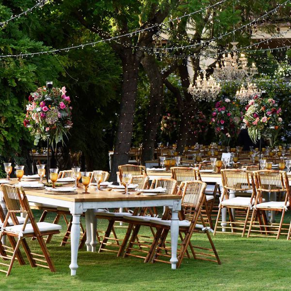 Montaje de mesas y sillas rústicas con decoración floral y luces colgantes para evento en jardín, organizado por Antonio Bort en Espartinas, Sevilla.